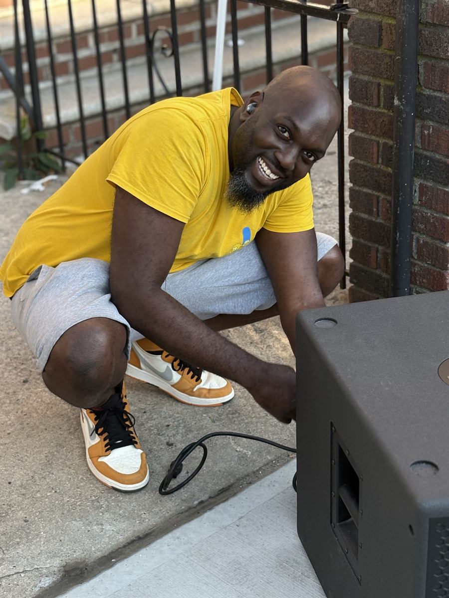 A neighbour in a yellow t-shirt crouched down on the sidewalk hooking up a speaker