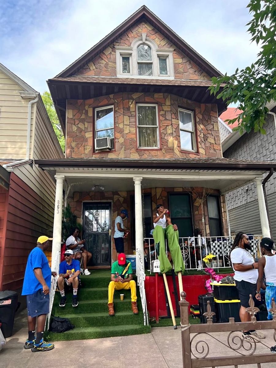 A stilt walker in green trousers towering over the front porch of 863 Albany Avenue while neighbors sit out on the steps