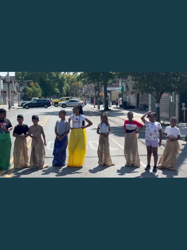 Nine kids lined up across the closed avenue standing in burlap sacks, waiting for the sack race to start