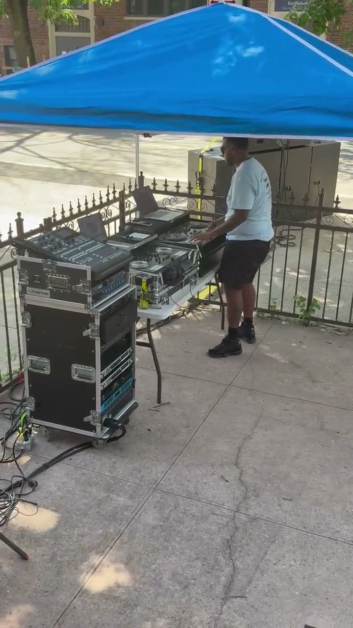 The DJ working the decks under a blue canopy on the sidewalk, speakers and road cases set up beside him