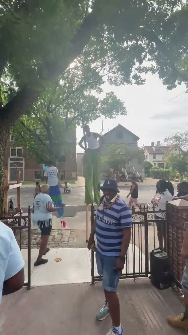 The stilt walker on tall green stilts making his way down the block while neighbors watch from the sidewalk and front steps